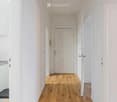 A narrow hallway with light wooden flooring and white walls, featuring a closed door at the end and an open door to the left leading to another room.