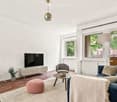 Modern living room with light-colored walls, wooden flooring, and large windows. Features a blue sofa, grey armchair, pink ottoman, coffee table, TV stand, and a small table with a potted plant.