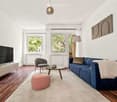 Modern living room with hardwood flooring, large windows, blue sofa, grey armchair, pink ottoman, coffee table, floor lamp, TV on side table, and decorative plant.