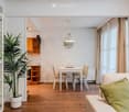 A modern, open-concept living and dining area with light wooden flooring, white walls, and large windows with beige curtains. Features include a white sofa with green cushions, a small dining table with four chairs, a potted plant, a wooden cabinet, and a