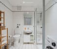 Modern bathroom with white tiles, a toilet, sink, mirror, shower, wooden shelf with towels, and a washing machine.
