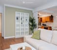 Bright living room with a white sofa, coffee table, bookshelf, and a potted plant. Adjacent kitchen area with wooden cabinets and a dining table.