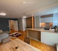 Open-plan living and dining area with a grey sofa, coffee table, dining table with chairs, and a modern kitchen island with cabinets, sink, and stove. The space features wooden flooring, recessed lighting, and a cowhide rug.