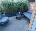 Balcony with a grey sofa, a small round table, and a wicker chair, surrounded by tall green plants.