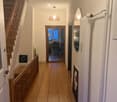 Wooden floor hallway with staircase on left, pendant light, mirror, radiator, and open doorway leading to another room with visible furniture.
