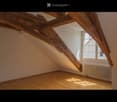Empty attic room with wooden beams, wooden floor, large window, radiator, and exposed wooden beams on the ceiling.