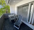 Balcony with dark tiled floor, white table, two chairs, and a lounge chair, with a view of greenery.