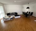 Modern living and dining area with light wooden flooring, white walls, and large windows with white curtains. Features a grey sectional sofa, white coffee table, and a black dining table with chairs.