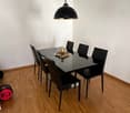 Dining room with a black glass table, six black chairs, and a hanging pendant light. A decorative candle is placed on the table.