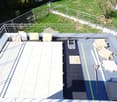 Roof terrace with metal railing, scattered construction materials including boards, tools, and a ladder, surrounded by green grass and trees.
