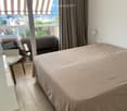 Modern bedroom with a large bed, a white chair, a small table with a lamp, and large windows with curtains offering a scenic view.