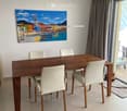 Modern dining room with a wooden dining table, four white chairs, and a large painting on the wall. The room features large windows with curtains and a view of the outside area.
