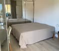 Modern bedroom with a large bed, a wardrobe with mirrored doors, a chair, a small table with a lamp, and a glass door leading to a balcony.