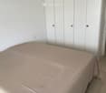 A modern bedroom with a neatly made bed covered in a light pink duvet, positioned against a white wall. A large white wardrobe with multiple doors is visible to the right of the bed.