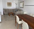 Modern living and dining area with a grey sectional sofa, wooden coffee table, dining table with white chairs, a mirror, and a floor lamp.