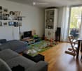 Living room with a grey sectional sofa, coffee table with books, a play mat with toys, a TV, a shelf with dishes, and a dining table with chairs. The room has wooden flooring and large windows with white curtains.
