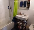 Modern bathroom with white tiles, a white sink, a bathtub, a toilet, and a shower area with a green curtain. Shelves hold towels and toiletries.