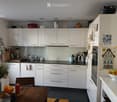 Modern kitchen with white cabinets, stainless steel appliances, and a wooden dining table with chairs. The counter features a sink, blender, and various kitchen items.