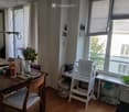 Bright dining area with wooden table, chairs, vase with flowers, and various items on the table. There is a shelf with a bowl and other small items near the window.