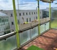 Balcony with wooden decking, green artificial grass, and several white planters with plants. The balcony has glass railings and is equipped with small lights.