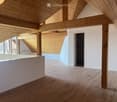 Empty room with wooden flooring, exposed wooden beams, and a white partition wall. The room has large windows and a doorway leading to another space.