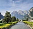 A scenic outdoor view featuring a river running through a valley with lush green grass on both sides, surrounded by towering mountains under a clear blue sky.