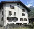 A two-story white house with brown shutters and a stone base, situated in a mountainous area with lush greenery and a clear blue sky.