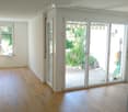 Empty room with light wooden flooring, white walls, large sliding glass doors leading to a garden, and a window on the left side.