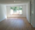 Empty room with light wooden flooring and white walls, featuring a large window with blinds and a door leading to another space.