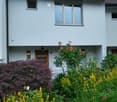 Front garden with colorful flowers, shrubs, and a small white bench near the entrance of a two-story house.
