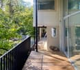 Wooden balcony with black railing, overlooking a lush green area with trees and a small stream. A table and chairs are placed on the balcony.