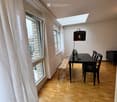 Bright, airy room with wooden flooring, large windows, a dining table with chairs, a bench, a sofa, and a floor lamp with a plant.