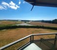 Balcony with metal railing overlooking a grassy field, road, and distant countryside with trees and mountains under a clear blue sky.