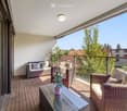 Balcony with wooden decking, wicker furniture including a sofa, chairs, and a glass coffee table with a flower pot, overlooking a green area with trees and buildings.