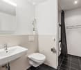 Modern bathroom with a white sink, chrome faucet, toilet, large mirror, and a washing machine in the adjacent space. The walls are tiled in light beige and black tiles, and a small flower vase is placed on the sink counter.