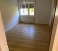 Empty room with light wooden flooring, white walls, and a window with blinds. A radiator is visible below the window.