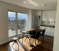Bright dining area with wooden table, chairs, and a stool, adjacent to a modern kitchen with white cabinets and a refrigerator, large windows offering a scenic view of rooftops and mountains.