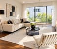 Modern living room with a beige sectional sofa, black and beige throw pillows, a glass coffee table with a black vase and books, a white leather chair, a floor lamp, a table lamp, and a large window with a view of greenery.
