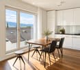 Modern kitchen and dining area with white cabinets, wooden table, black chairs, and a view of a tiled roof and mountains.