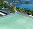 A modern outdoor pool area with a panoramic view of a lake and surrounding mountains, featuring potted plants and a glass railing.