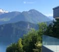 Modern building with a glass balcony overlooking a lake and snow-capped mountains.
