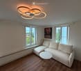 Modern living room with light-colored sofa, round marble coffee table, large windows, and a contemporary ceiling light fixture.