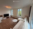 Modern living room with wooden flooring, large windows, a beige sofa, a marble coffee table, a flat-screen TV on a wooden stand, and a wall-mounted cabinet.