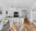 Open-plan living and dining area with a grey sofa, coffee table, bar stools, kitchen island, and modern appliances. The space features pendant lights, framed pictures, and a TV stand.