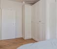 Minimalist bedroom with light-colored wooden flooring, white walls, a single bed with light blue bedding, and built-in white wardrobes.