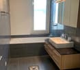 Modern bathroom with dark tiled walls and floor, a bathtub, a sink with a wooden cabinet, and a toilet. Features include a large window, built-in lighting, and a stylish mirror cabinet.