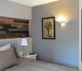 Modern bedroom with a grey bed, nightstand, lamp, mirror, and a picture frame on the wall. The room features a wooden headboard and a built-in shelf above the bed.