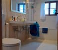Modern bathroom with a toilet, double sink, bathtub, and large mirror. Features tiled walls, wooden accents, and a window with a curtain.