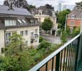 View of a multi-story residential building with solar panels on the roof, surrounded by greenery and other houses in the background.