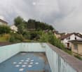 An abandoned rooftop area with a blue tiled floor and light blue walls, surrounded by residential houses and greenery.